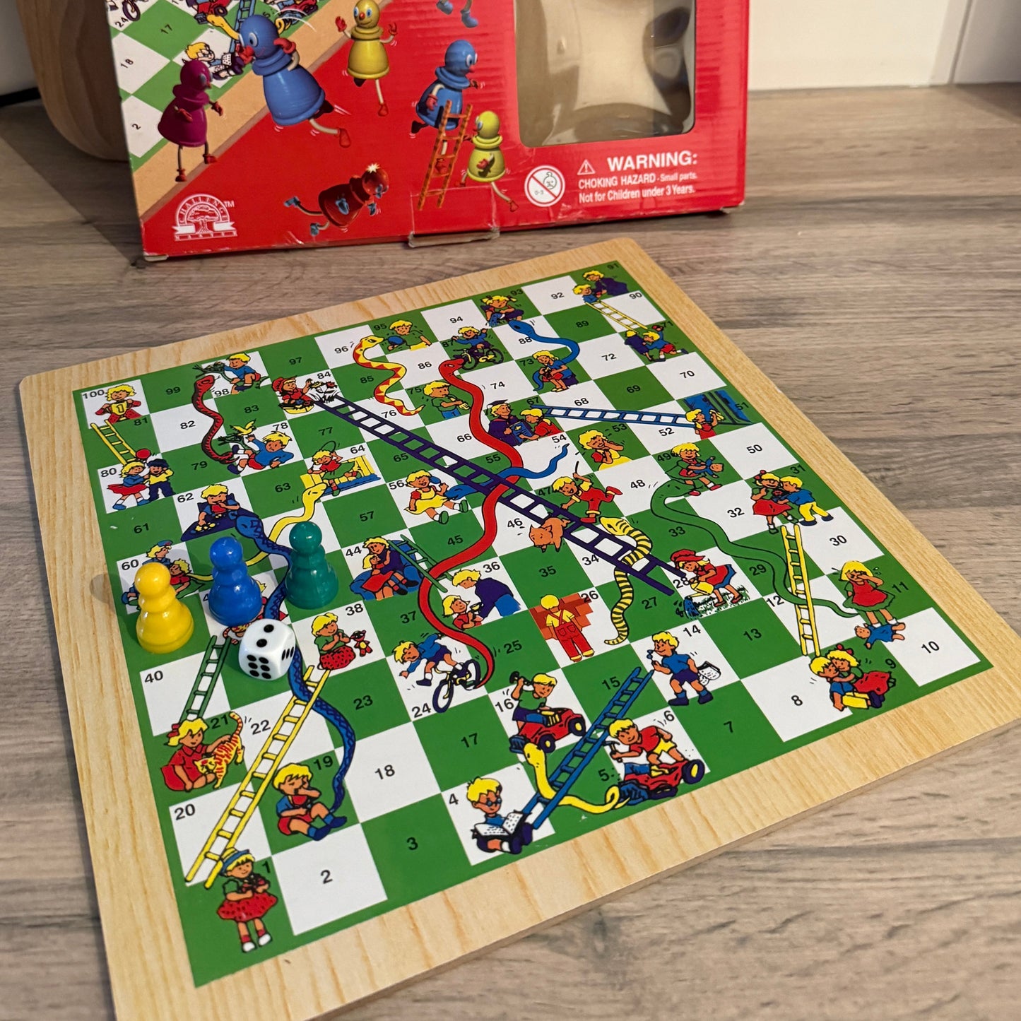 Children's Snakes And Ladders board game with colorful pieces on a wooden floor, next to its packaging.